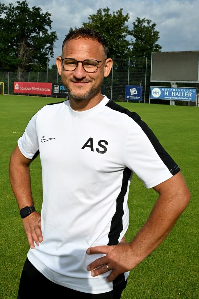 Ein Mann mit Brille und einem weißen AS-Sportshirt steht lächelnd mit den Händen in den Hüften auf einem grünen Fußballplatz