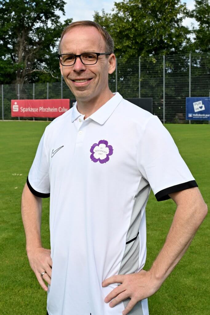 Ein Mann mit Brille und weißem Polo steht lächelnd mit den Händen in den Hüften auf einer Wiese, im Hintergrund sind Bäume und Schilder zu sehen