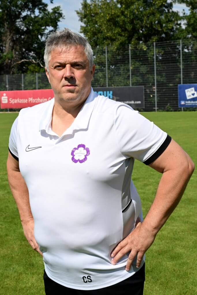 Ein grauhaariger Mann in einem weißen Poloshirt steht mit den Händen in den Hüften auf einem grünen Fußballfeld und blickt in die Kamera