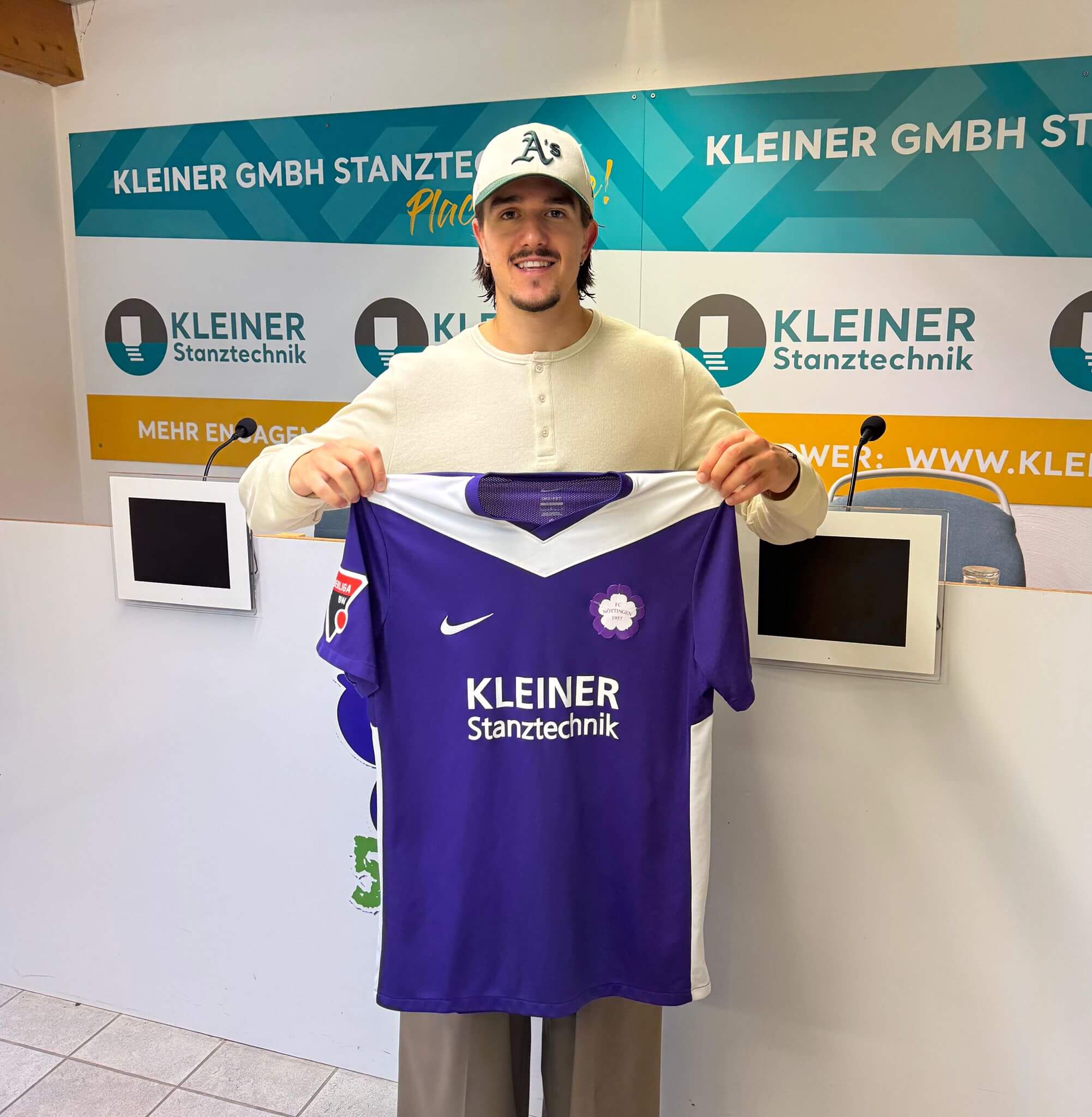 Antonio Babic mit weißer Mütze präsentiert ein lila FCN-Trikot vor dem Schild von „Kleiner Stanztechnik“