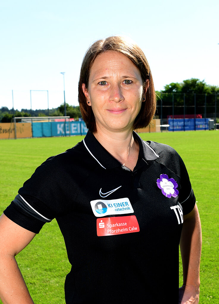 Eine Frau in einem schwarzen Poloshirt steht auf dem sonnigen Fußballplatz und blickt als Juniorenleiterin neutral in die Kamera