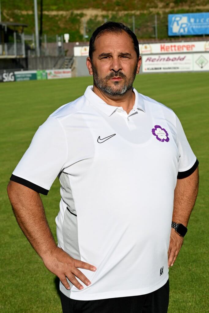 Ein Mann in einem weißen Trikot steht mit den Händen in den Hüften auf einem grünen Feld und schaut in die Kamera