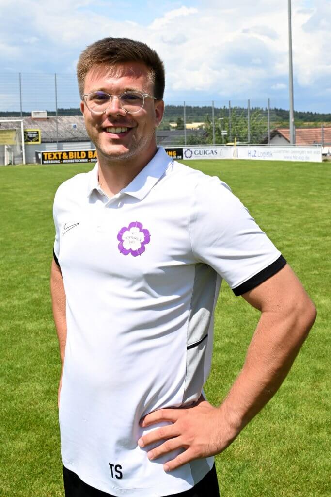 Ein lächelnder Mann mit Brille und weißem Polohemd steht auf einem grünen Fußballplatz und blickt mit einer Hand in die Hüfte zur Kamera