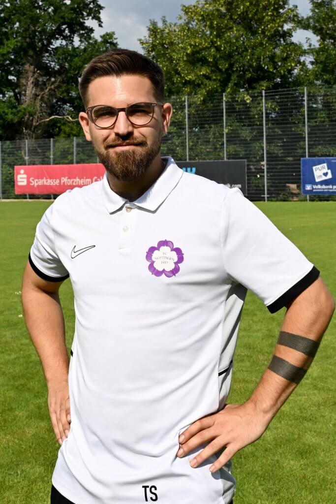 Ein Mann mit Brille und weißem Poloshirt steht auf einer Wiese, die Hände in die Hüften gestützt, und schaut in die Kamera