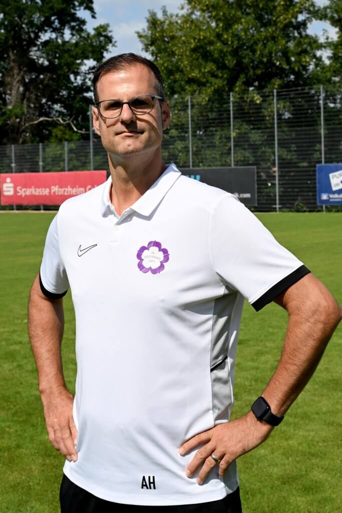 Ein Mann in einem weißen Poloshirt steht mit den Händen in den Hüften auf einer Wiese, trägt eine Brille und eine Uhr