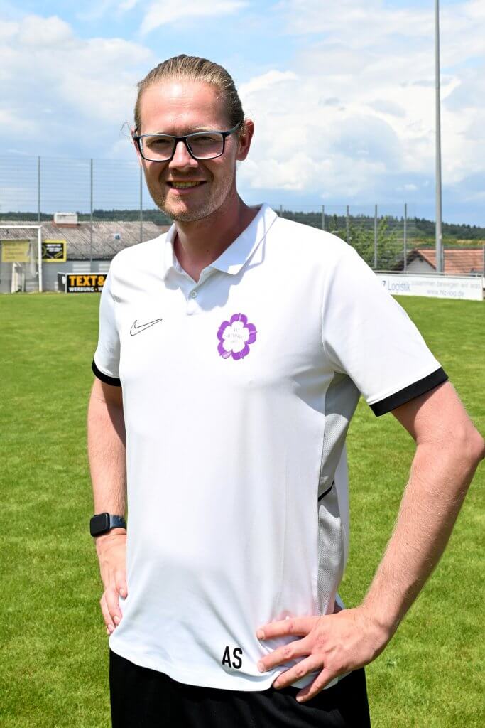 Eine lächelnde Person mit Brille in einem weißen Trikot steht auf einem grünen Fußballfeld, im Hintergrund sind Schilder und Bäume zu sehen