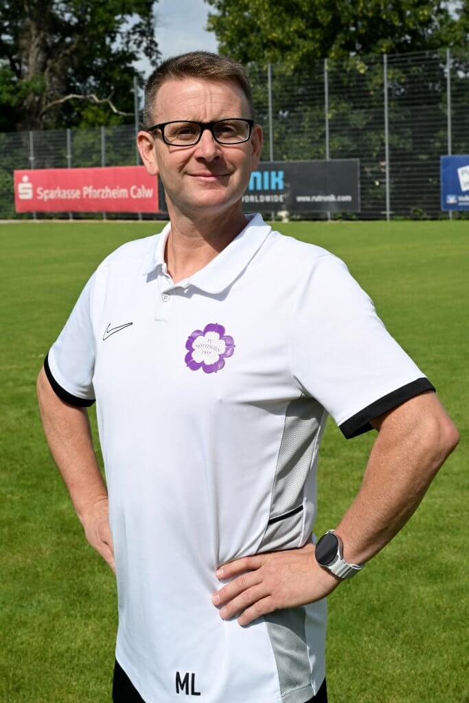 Ein Mann mit Brille und weißem Poloshirt steht auf einem Fußballfeld, die Hände in die Hüften gestützt, und blickt in die Kamera