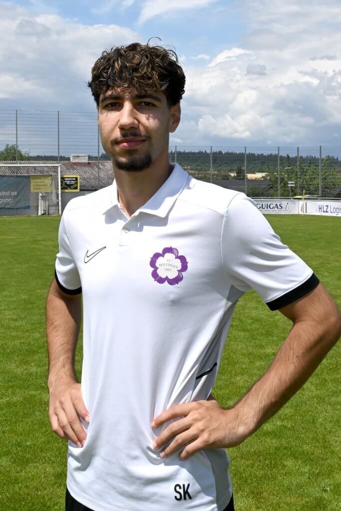 Ein junger Mann in einem weißen Trikot steht mit den Händen in den Hüften auf dem Rasen und schaut direkt in die Kamera