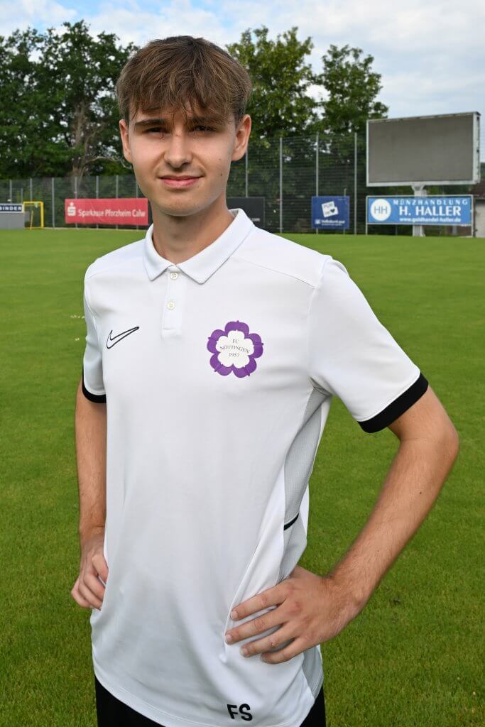 Ein junger Mann in einem weißen Trikot steht lächelnd mit den Händen in den Hüften auf einem Sportplatz, im Hintergrund sind Bäume und Schilder zu sehen