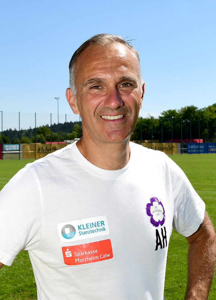 Ein lächelnder Mann in einem weißen Hemd steht auf einem Sportplatz mit Junioren-Toren und Bäumen im Hintergrund