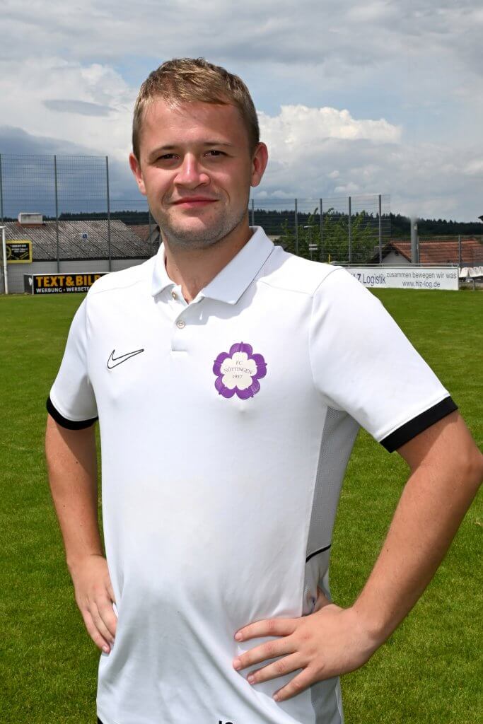 Ein Mann in einem weißen Polohemd steht lächelnd mit den Händen in den Hüften auf einem Fußballplatz