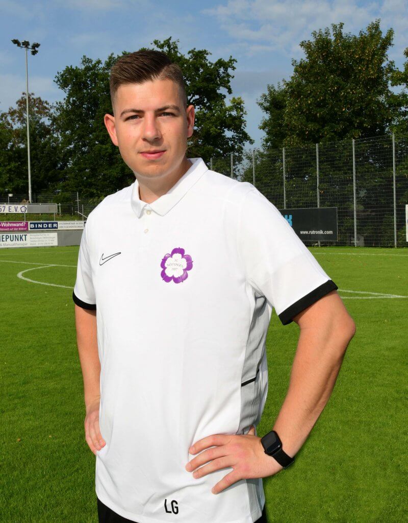 Eine Person in einem weißen Poloshirt steht mit den Händen in den Hüften auf dem Fußballfeld und schaut in die Kamera