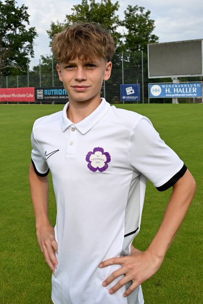 Ein Jugendlicher in einem weißen Fußballtrikot steht mit einer Hand in der Hüfte auf dem Spielfeld und blickt in die Kamera