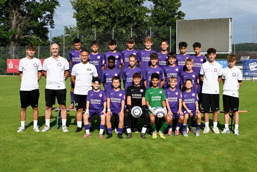 Eine Jungenfußballmannschaft mit Trainern steht auf einem Rasen, alle in lila Trikots; zwei von ihnen halten vorne einen Fußball in den Händen.