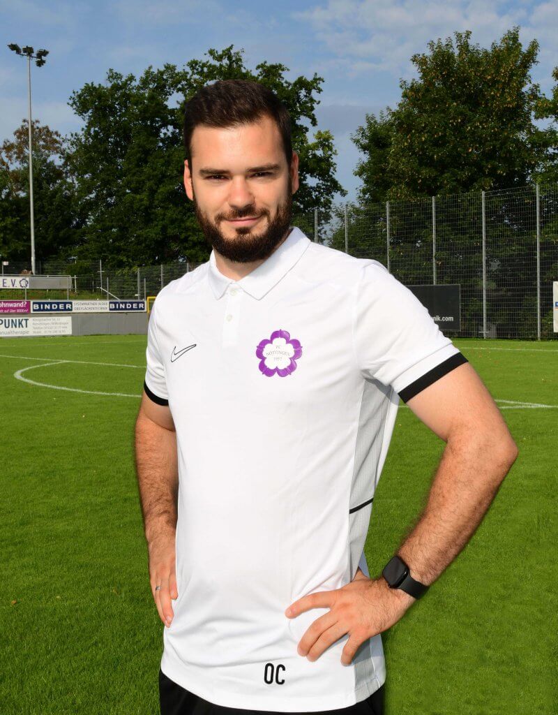 Ein Mann in einem weißen Polohemd mit Blumenlogo steht lächelnd auf einem Fußballplatz und hat die Hände in die Hüften gestemmt