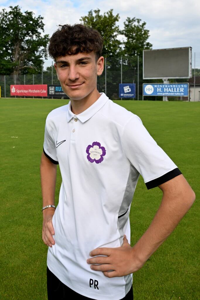 Ein Jugendlicher in einem weißen Polohemd steht lächelnd mit den Händen in den Hüften auf einem Fußballfeld, im Hintergrund sind Bäume und Schilder zu sehen