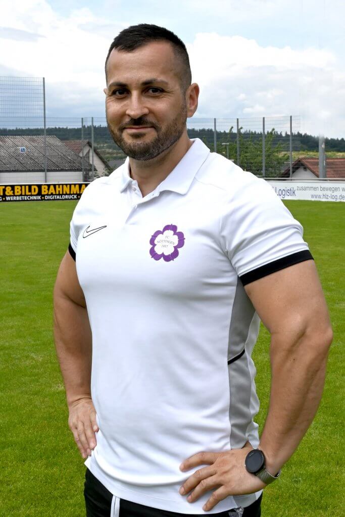 Ein Mann in einem weißen Polohemd steht mit einer Hüftstütze auf dem Fußballplatz und blickt als Juniorenleiter in die Kamera