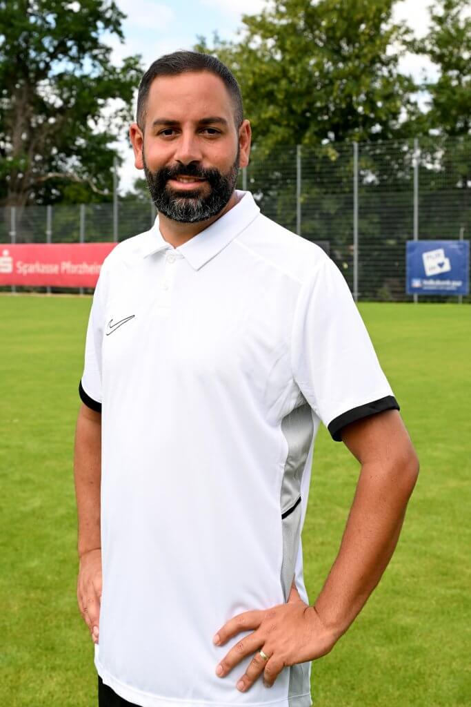 Ein Mann in einem weißen Poloshirt steht auf einer Wiese, im Hintergrund sind Bäume und Zäune zu sehen