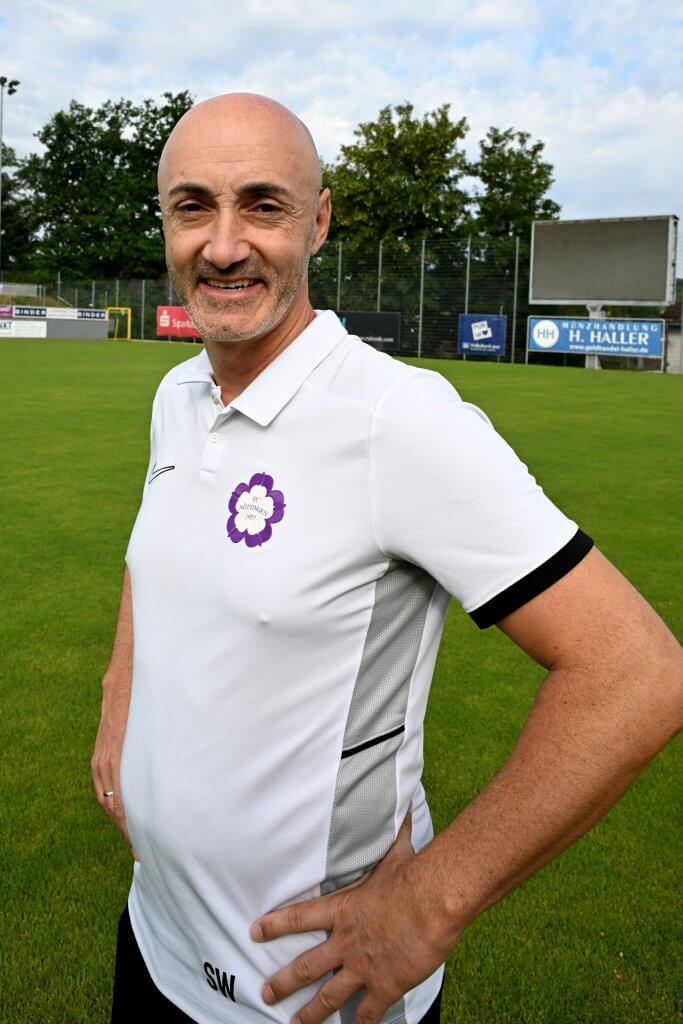Ein lächelnder, kahlköpfiger Mann in einem weißen Polohemd steht auf einem Fußballplatz, im Hintergrund sind Bäume und Werbebanner zu sehen