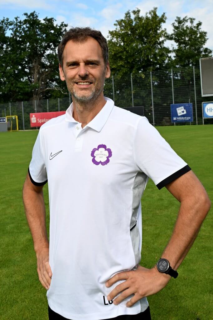 Ein Mann in einem weißen Sportpolo steht auf einer Wiese, lächelt, hat die Hände in die Hüften gestemmt, und im Hintergrund sind Bäume zu sehen