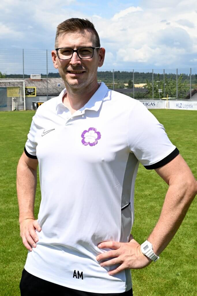 Ein Mann mit Brille und weißem Polohemd aus der Juniorenleitung steht lächelnd mit den Händen in den Hüften auf dem Fußballfeld