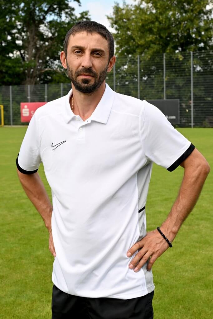 Ein Mann in einem weißen Polohemd steht mit den Händen in die Hüften gestützt auf einer Wiese, im Hintergrund sind Bäume und ein Zaun zu sehen
