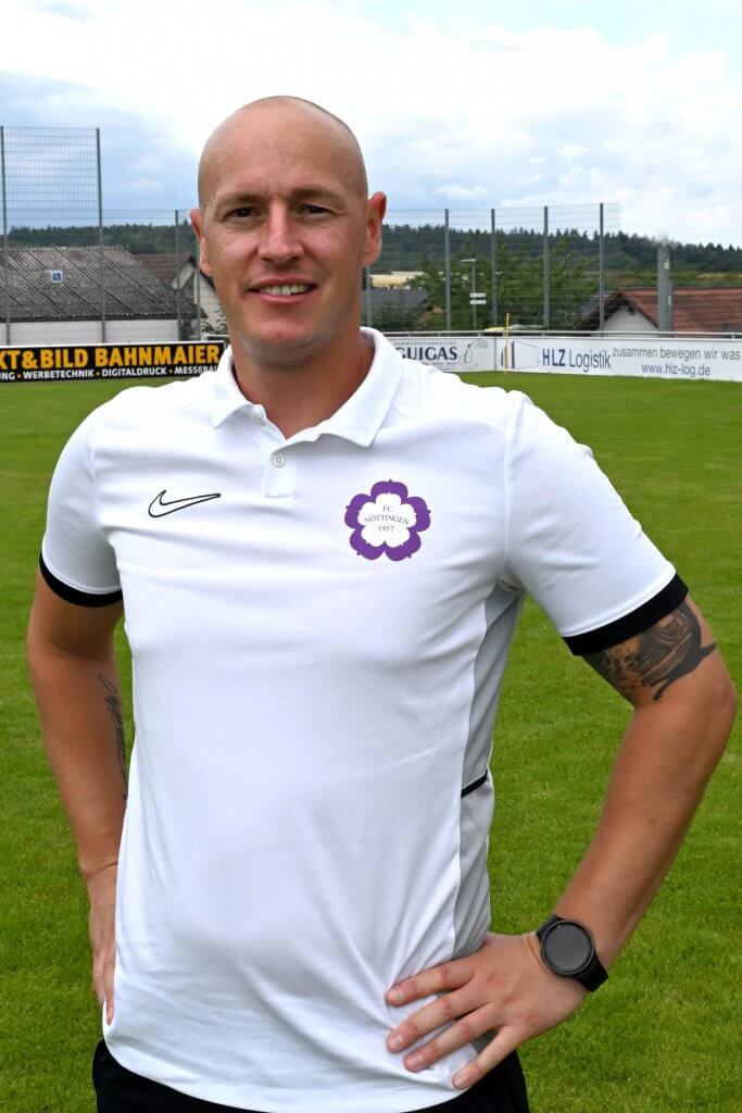Ein Mann mit Glatze in einem weißen Poloshirt steht lächelnd mit den Händen in den Hüften auf einem Fußballfeld